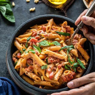 tomato basil pasta in a bowl with a fork digging in into the bowl. There are also some basil leaves and a small jar of olive oil in the background