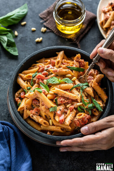 tomato basil pasta in a bowl with a fork digging in into the bowl. There are also some basil leaves and a small jar of olive oil in the background