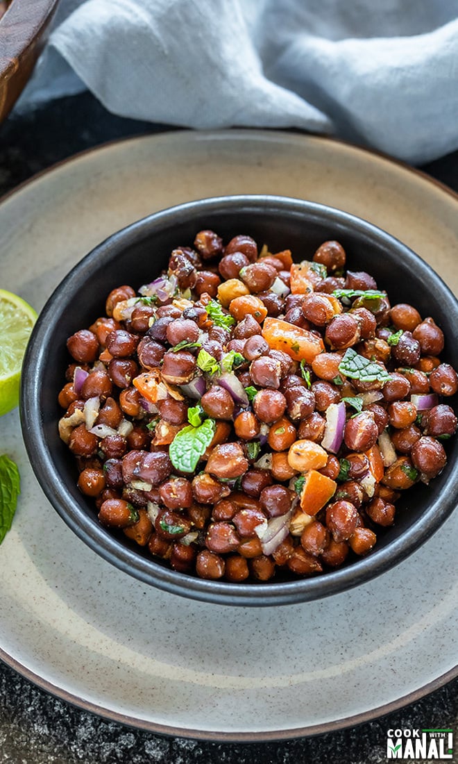 a black bowl full of kala chana chaat with a lime wedge and mint leaf on the side