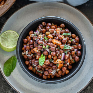 a black bowl full of kala chana chaat with a lime wedge and mint leaf on the side
