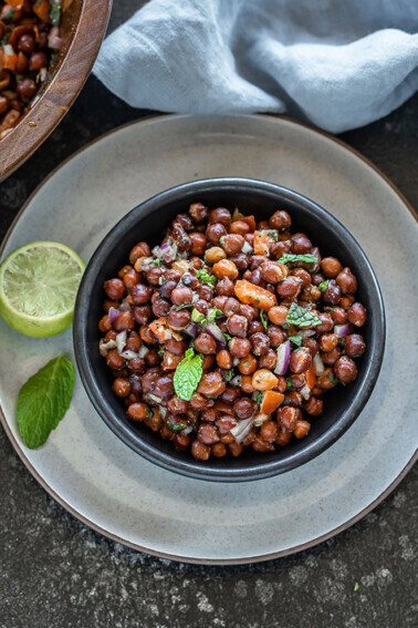 a black bowl full of kala chana chaat with a lime wedge and mint leaf on the side