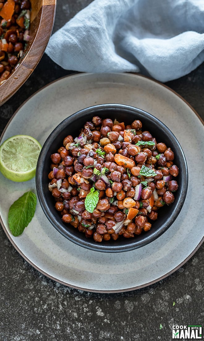 a black bowl full of kala chana chaat with a lime wedge and mint leaf on the side