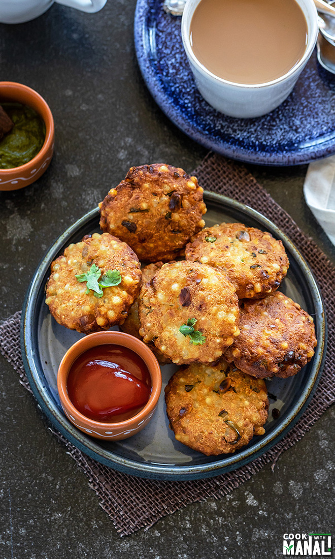 sabudana vada in a plate served with a small bowl of tomato ketchup and a cup of chai in the background