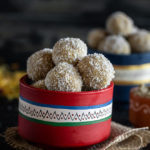 thandai coconut ladoo in a red color bowl with more ladoos and some flowers in the background
