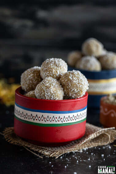 thandai coconut ladoo in a red color bowl with more ladoos and some flowers in the background