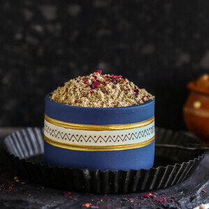 thandai powder in a blue color bowl with rose petals scattered on the sides