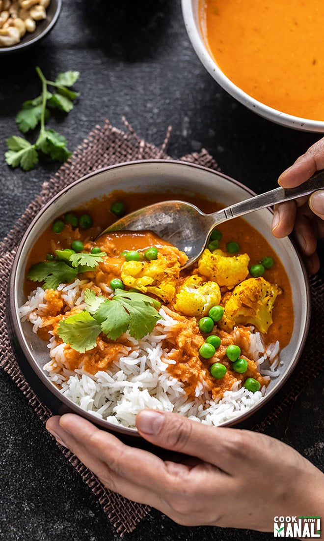 pair of hands holding a bowl of curry and rice from one side and digging into the bowl with a spoon from another hand