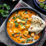 bowl of paneer butter masala served with a piece of naan and garnished with cilantro and cream