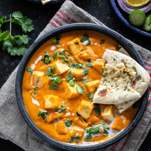 bowl of paneer butter masala served with a piece of naan and garnished with cilantro and cream
