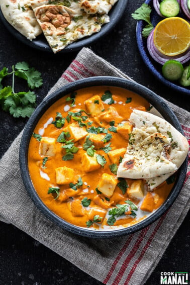 bowl of paneer butter masala served with a piece of naan and garnished with cilantro and cream