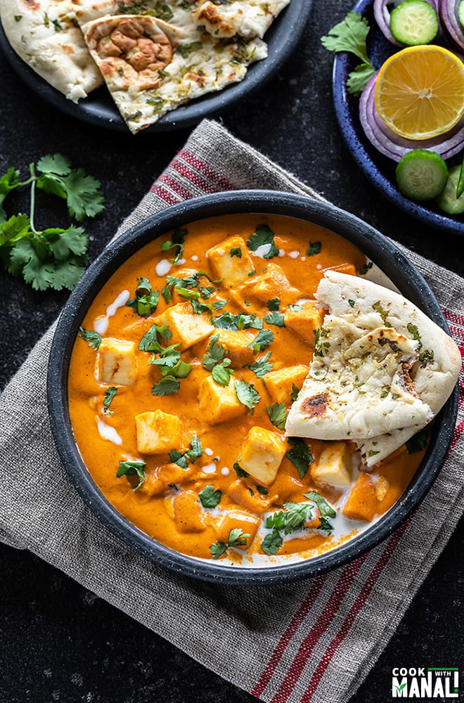 bowl of paneer butter masala served with a piece of naan and garnished with cilantro and cream