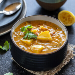 potato curry served in a black bowl, garnished with cilantro with a bowl of rice and a squeezed lemon placed in the background