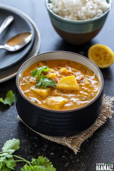 potato curry served in a black bowl, garnished with cilantro with a bowl of rice and a squeezed lemon placed in the background