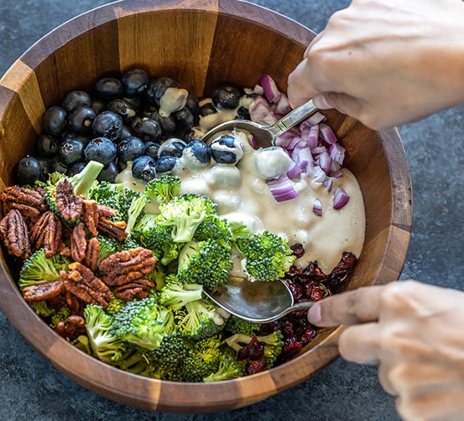 a bowl of vegan broccoli salad being mixed with the dressing using 2 spoons