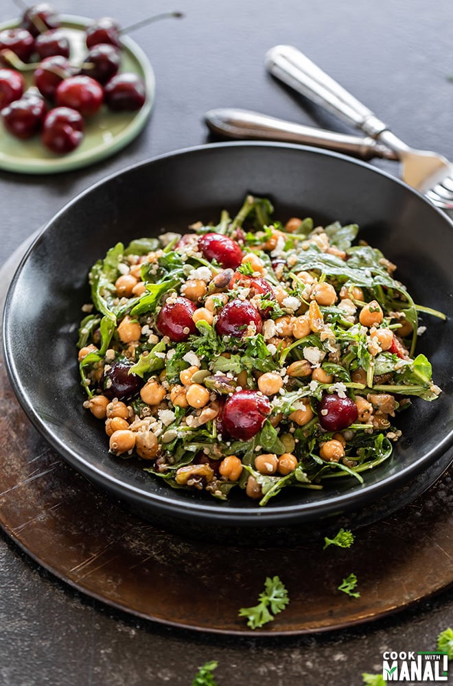 black color salad bowl filled with cherries, quinoa, arugula, chickpeas, pistachios with more cherries in the background