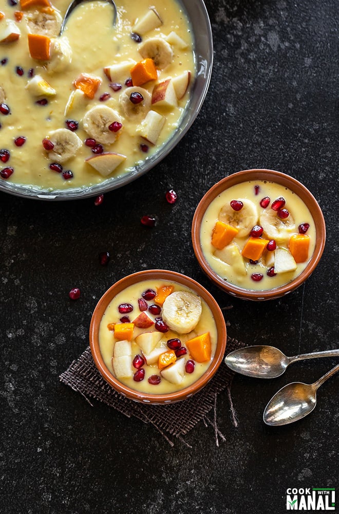 two clay bowls filled with fruits custard with spoons on the side and a large bowl of fruit custard in the background