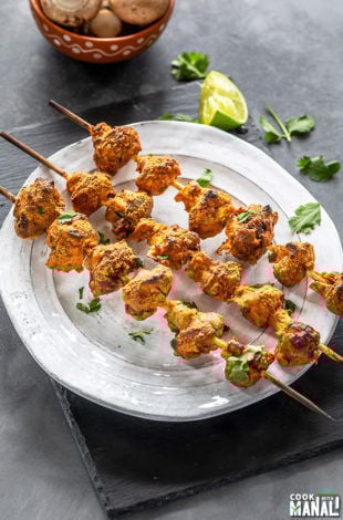 three wooden skewers of mushroom tikka arranged on a white plate and garnished with cilantro