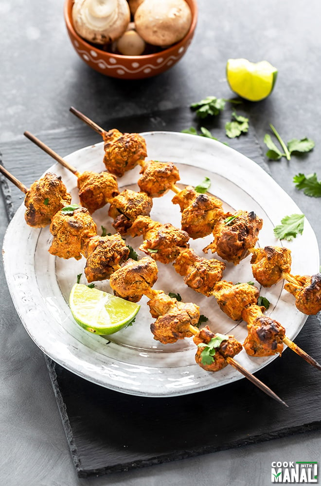 three wooden skewers of mushroom tikka arranged on a white plate and garnished with cilantro