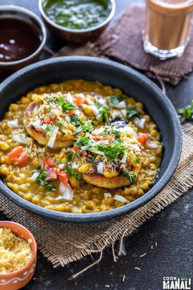 ragda patties served in a black bowl with a cup of chai and several bowls of chutney in the background and on the sides