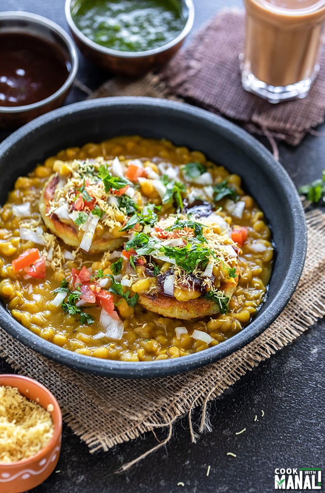 ragda patties geserveerd in een zwarte kom met een kop chai en diverse kommen chutney op de achtergrond en aan de zijkanten