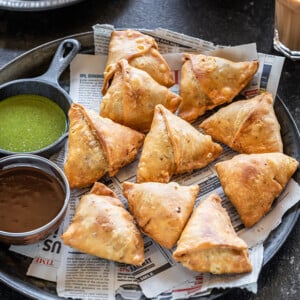 samosa served in a plate with two bowls of chutney and glass of chai on the side