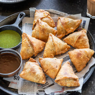 samosa served in a plate with two bowls of chutney and glass of chai on the side