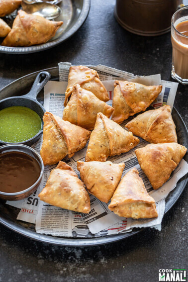 samosa served in a plate with two bowls of chutney and glass of chai on the side