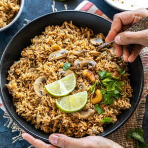 pair of hand holding a bowl of mushroom biryani with one hand digging into the bowl with a spoon