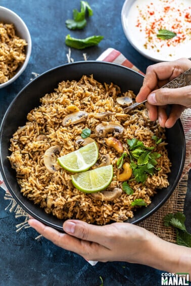 pair of hand holding a bowl of mushroom biryani with one hand digging into the bowl with a spoon