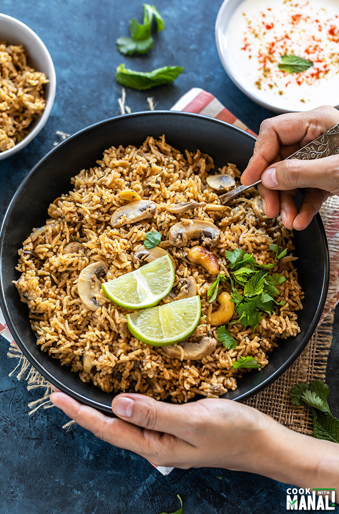 pair of hand holding a bowl of mushroom biryani with one hand digging into the bowl with a spoon