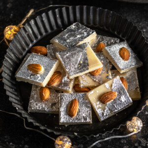 badam burfi placed in a black plate surrounded by lights and a plate of almonds in the background