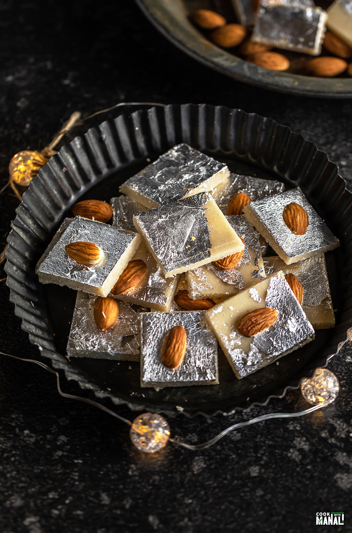 badam burfi placed in a black plate surrounded by lights and a plate of almonds in the background