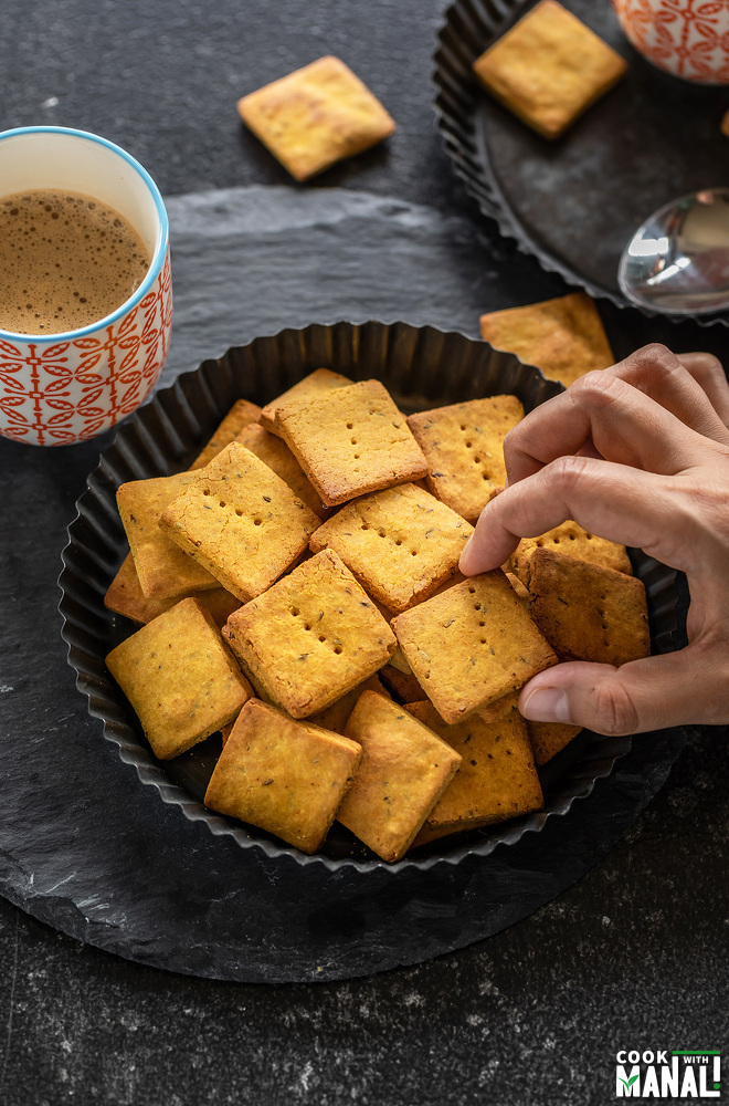 a hand picking up besan papdi from a plate with a cup of coffee in the background