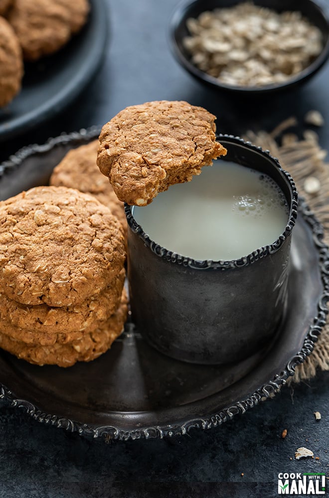 half eaten cookie placed on side of a cup of milk and more oats jaggery cookies in the background