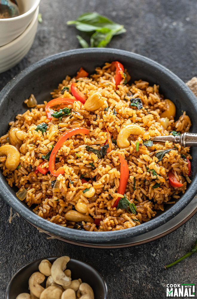 thai basil fried rice in a black bowl with a fork with a small bowl of cashews on the side and few basil leaves scattered around