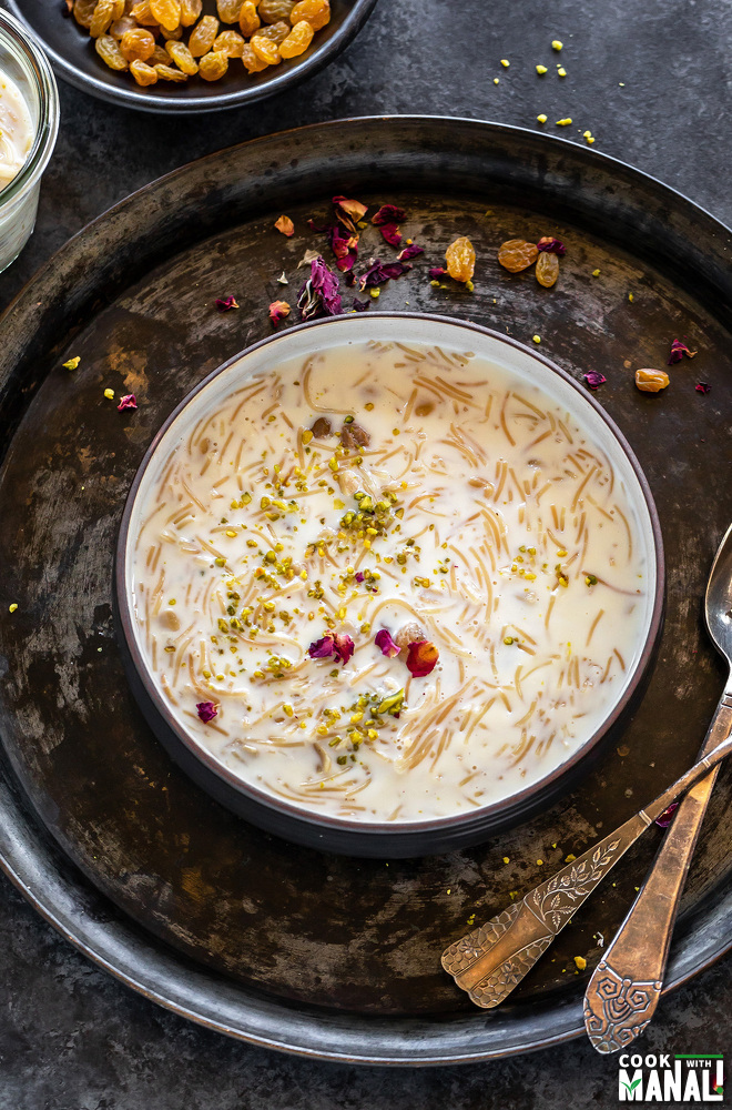 seviyan kheer served in a black bowl garnished with rose petals and pistachios with more rose petals and nuts scattered in the background