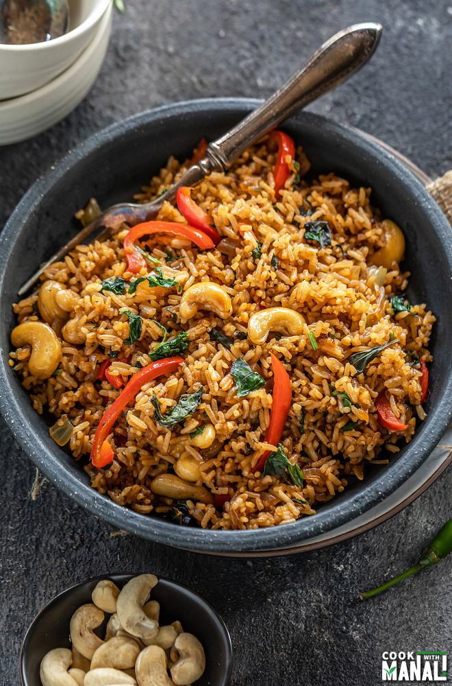 thai basil fried rice in a black bowl with a fork with a small bowl of cashews on the side and few basil leaves scattered around