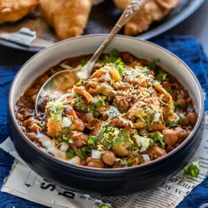 samosa chaat in a black bowl with a spoon with plate full of samosa in the background