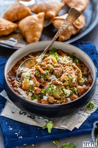 samosa chaat in a black bowl with a spoon with plate full of samosa in the background