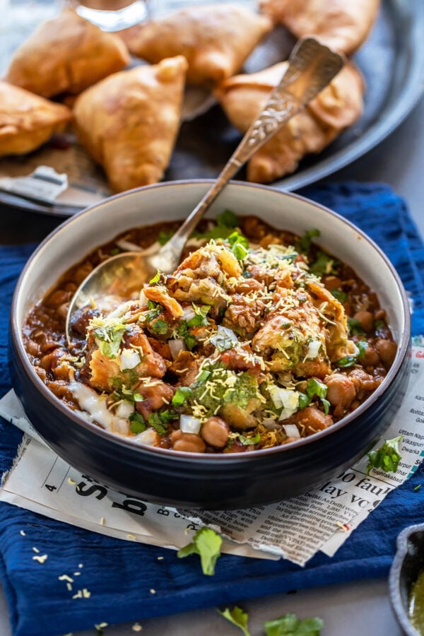 samosa chaat in a black bowl with a spoon with plate full of samosa in the background