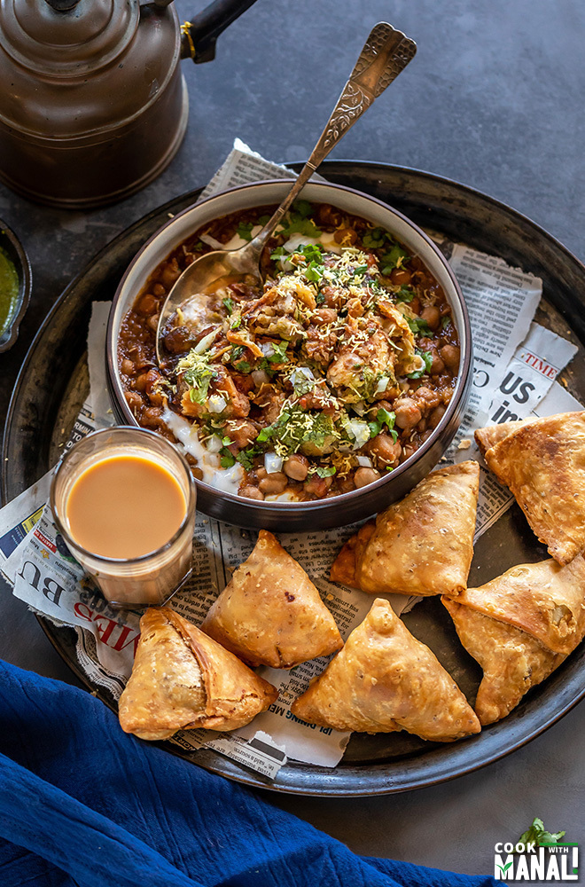 large round plate with bowl of samosa chaat, few samosa on the side and a cup of chai with a kettle in the background