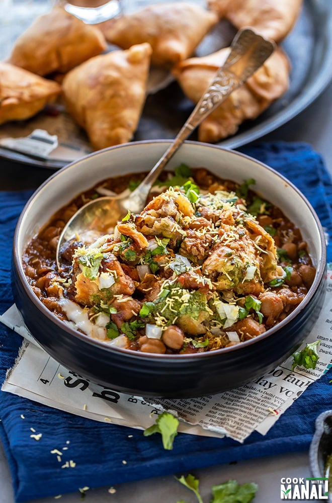 samosa chaat in a black bowl with a spoon with plate full of samosa in the background