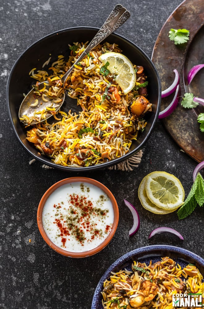 a bowl of vegetable biryani with a spoon placed along a bowl of yogurt and some lemon wedges and sliced onion scattered all around