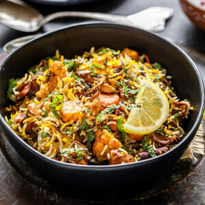 bowl of vegetable biryani garnished with lemon wedge and some spoons placed in the background along with a plate of sliced onions