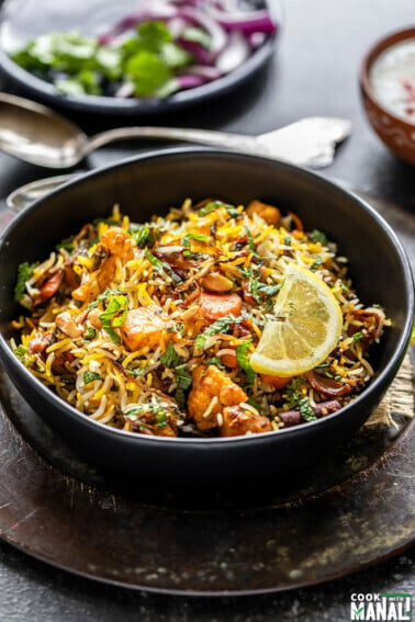 bowl of vegetable biryani garnished with lemon wedge and some spoons placed in the background along with a plate of sliced onions