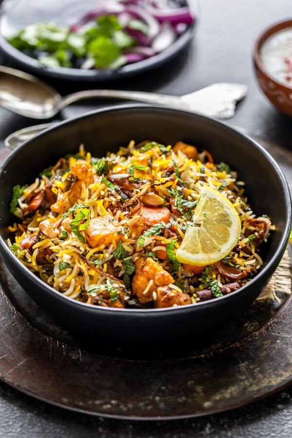 bowl of vegetable biryani garnished with lemon wedge and some spoons placed in the background along with a plate of sliced onions