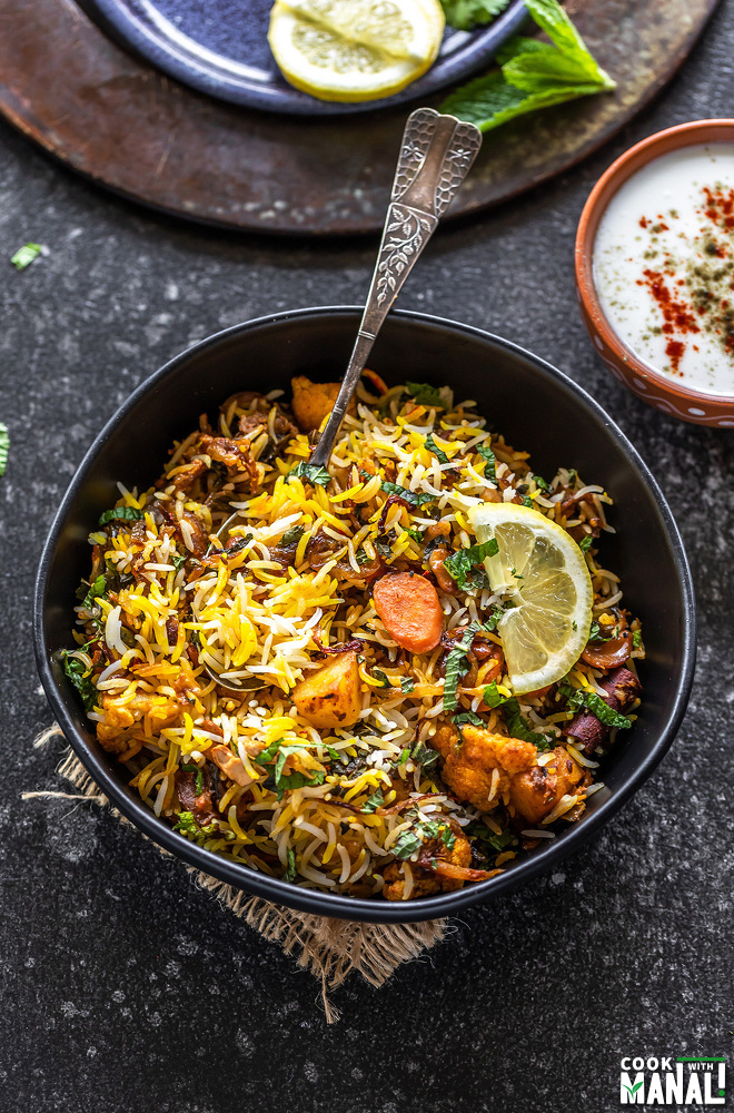 bowl of vegetable biryani with a spoon and a bowl of yogurt placed on the side