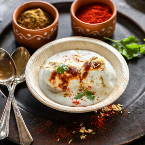 dahi bhalla served in a bowl and garnished with chutneys with two spoons kept on on side and bowls of spices in the background
