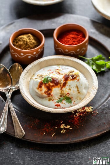 dahi bhalla served in a bowl and garnished with chutneys with two spoons kept on on side and bowls of spices in the background