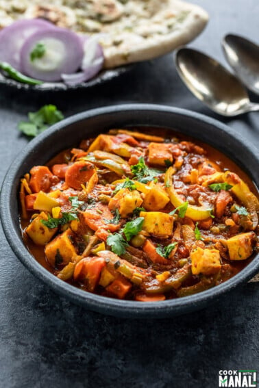 vegetable jalfrezi served in a black bowl with naan placed in the background along with some onion slices